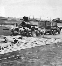 Soldiers of IX Engineering Command, U.S. Army Air Force, putting down a Pierced Steel Planking (PSP) Runway at an Advanced Landing Ground under construction somewhere in France following the Normandy Landings of World War II